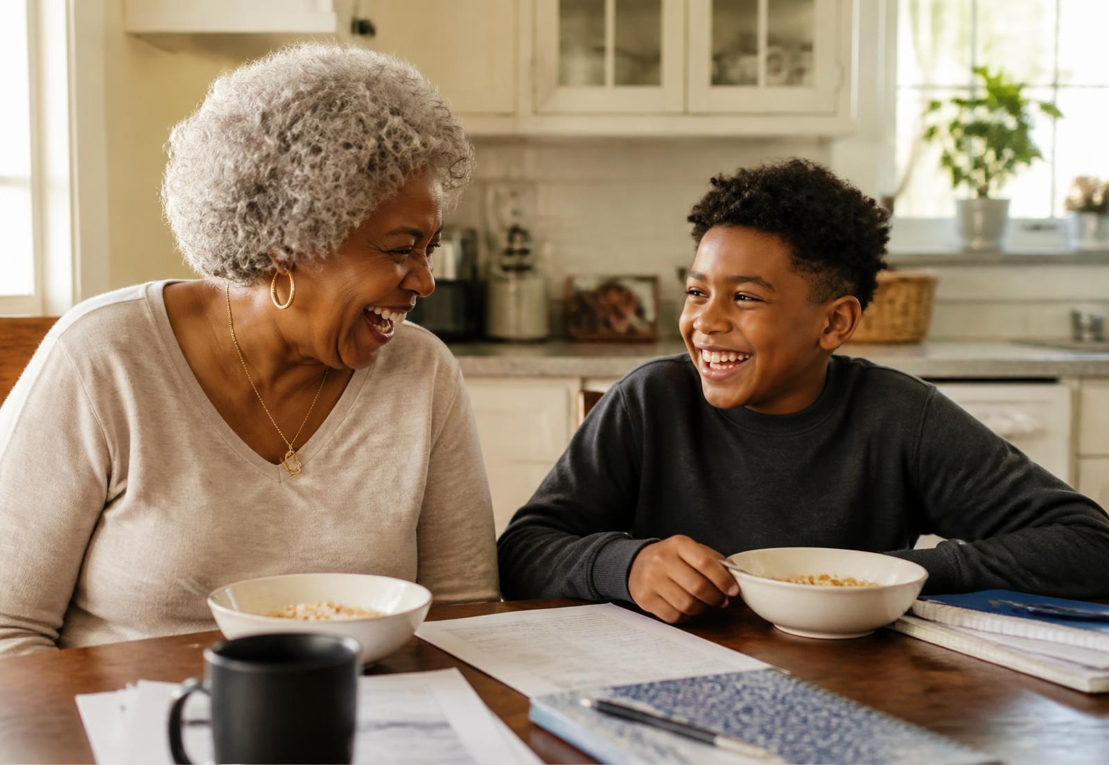A grandmother with silver curls and her grandson laughing together at a kitchen table over cereal and homework papers.