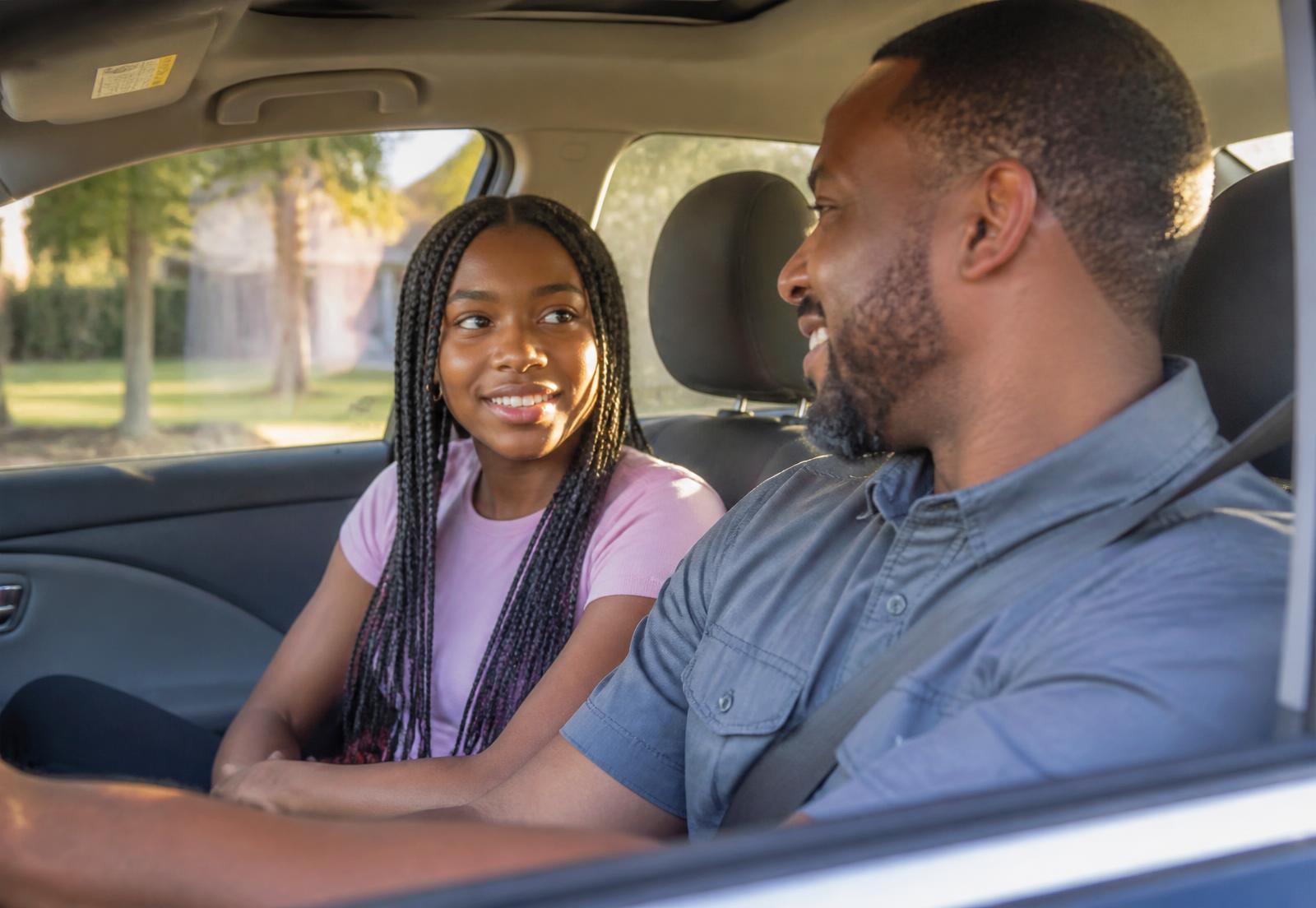 A father and his teenage daughter smiling at each other in a parked car filled with warm afternoon sunlight.