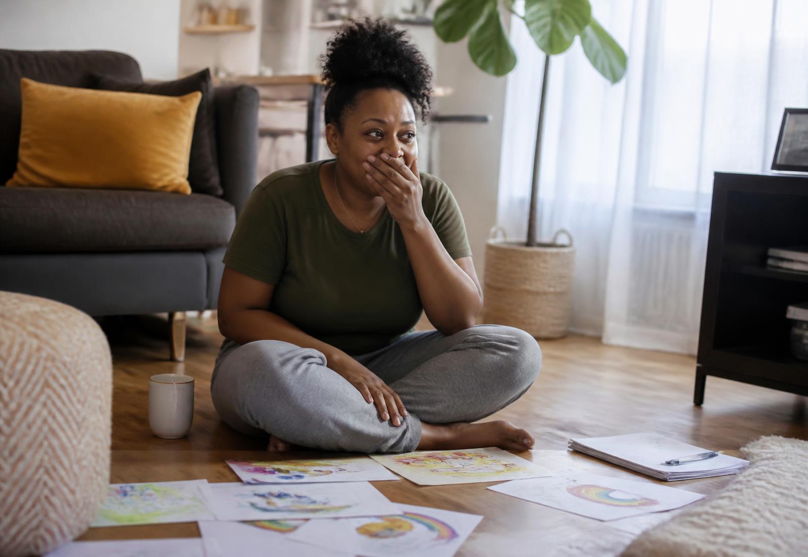 A mother sitting on the living room floor with her child's drawings, hand over her mouth in a moment of relief and recognition.